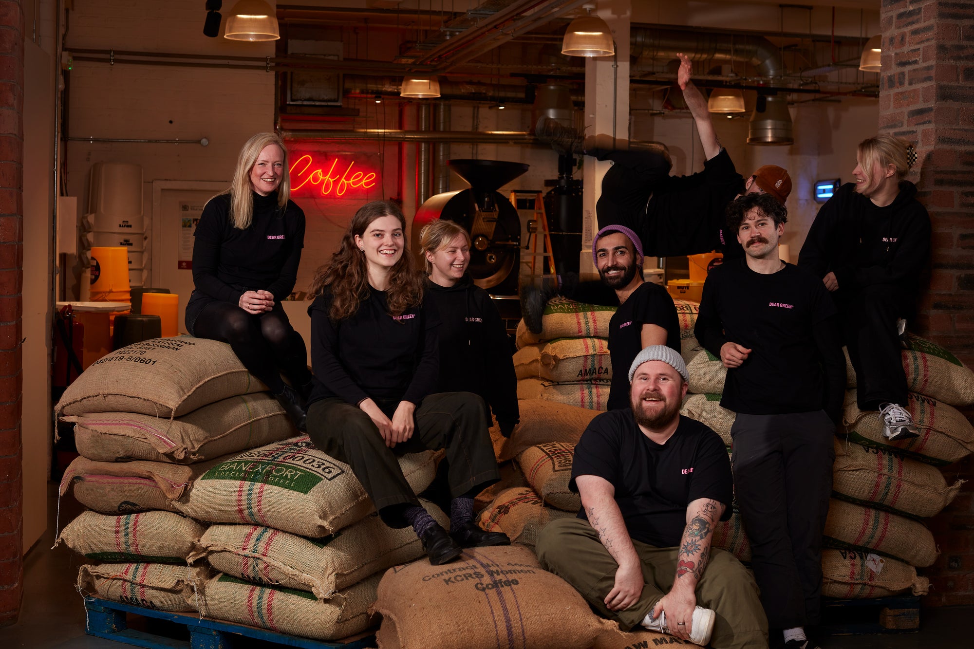 The staff members of Dear Green sitting on bags of raw green coffee beans. There are 7 staff members in the photo, all wearing black Dear Green tops and smiling at the camera.