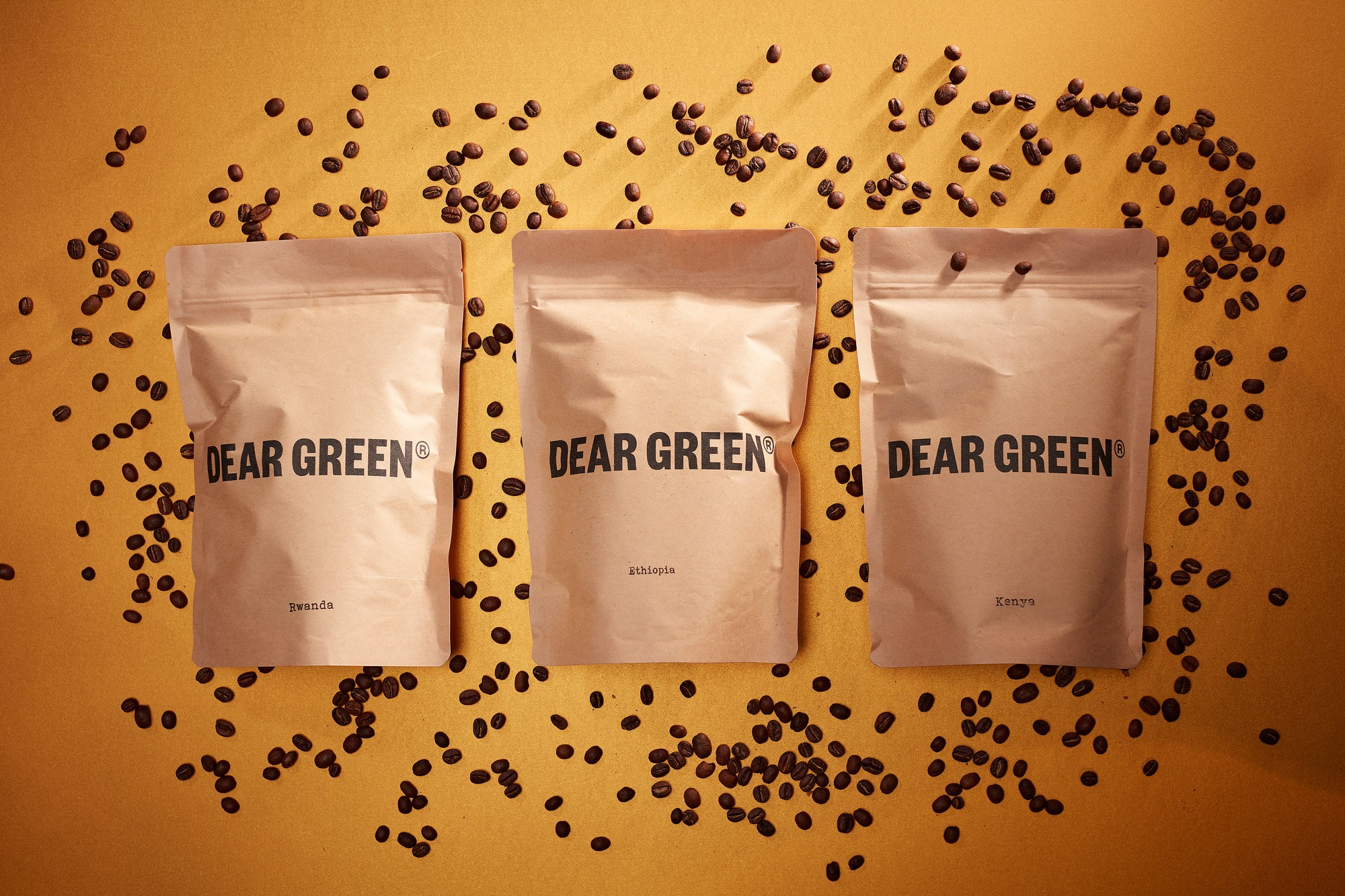 Three brown bags of coffee beans with the Dear Green logo, each labeled with their origin - Rwanda, Ethiopia and Kenya. Scattered around the bags are loose coffee beans and the composition is on a warm yellow background.