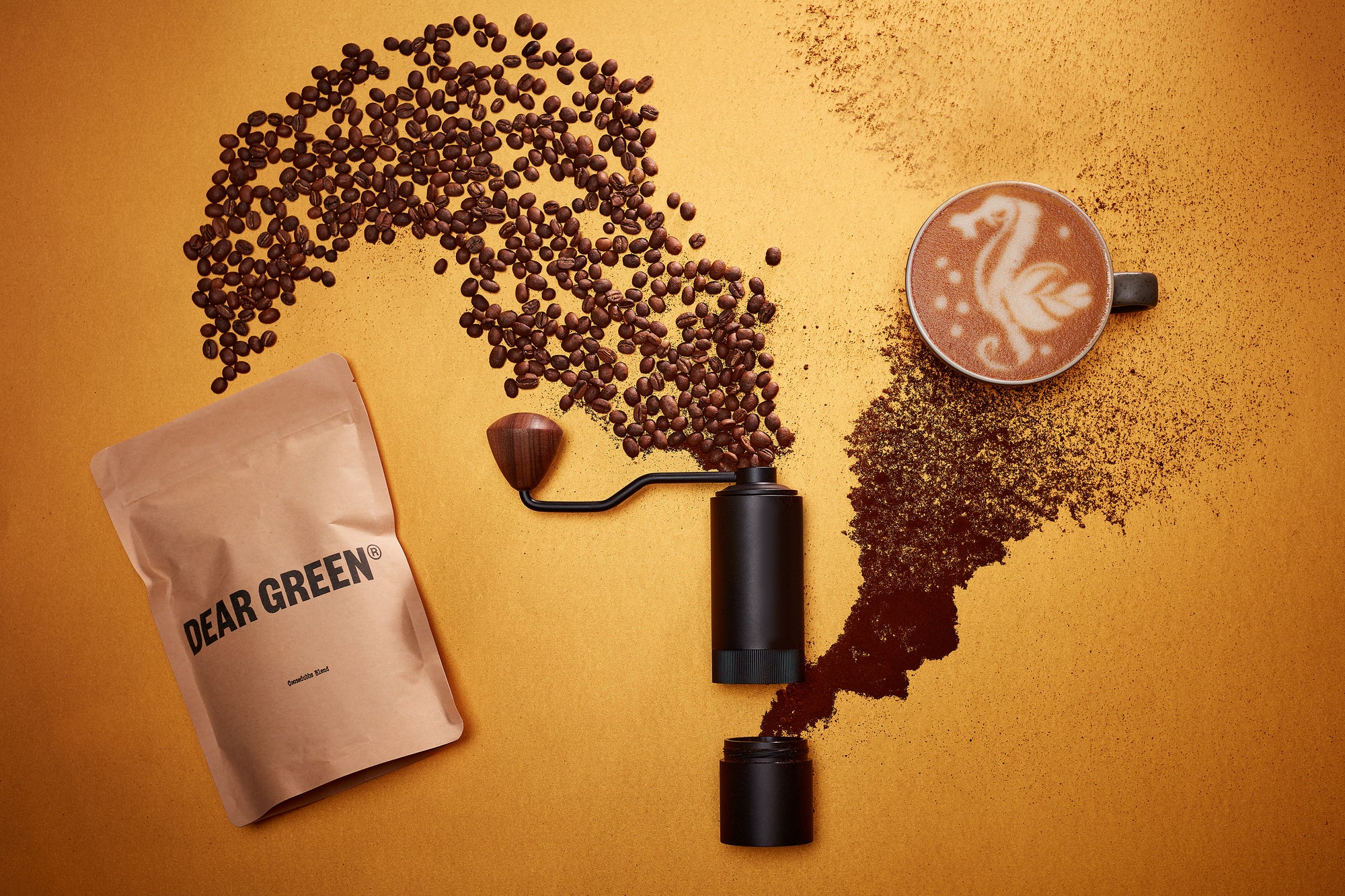 Flatlay of a bag of Dear Green Coffee beans with loose beans scattered decoratively alongside a coffee grinder and a latte with dragon latte art. The composition is on a warm yellow background.