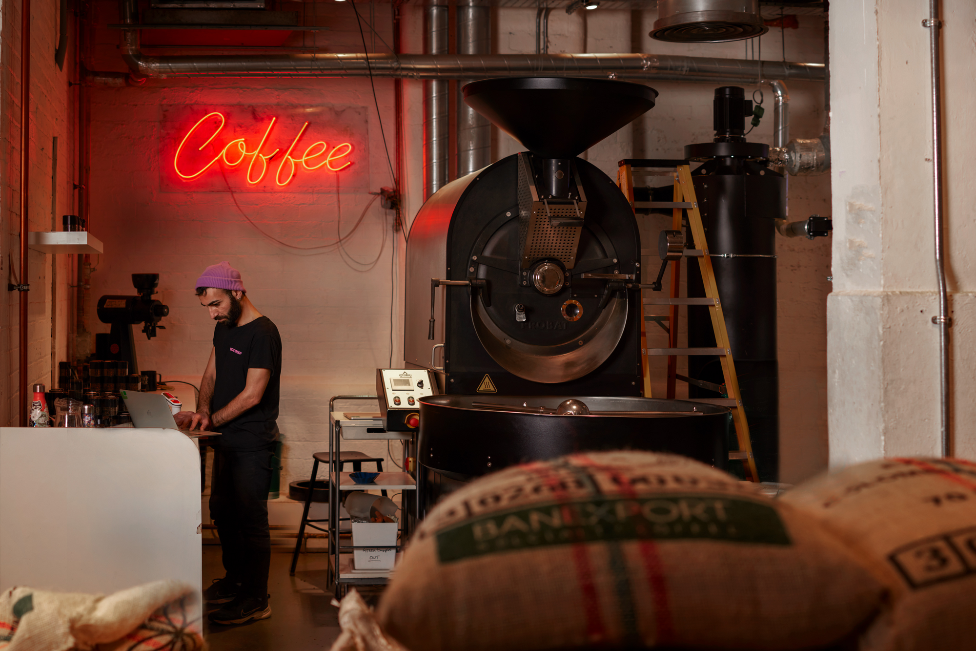 Roastery in action with an illuminated "coffee" sign and coffee roaster standing in the roasting area with bags of beans in the foreground. The staff member is wearing black with a purple beanie and is deep in concentration.